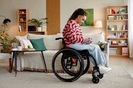 Young adult Caucasian woman with disability sitting in wheelchair using digital tablet in living room, focusing on screen, long hair styled in braids, casual home environment