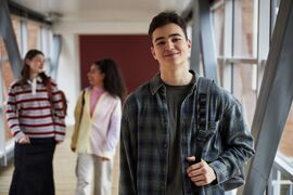 Portrait of Caucasian teenage boy smiling at camera while holding backpack in university hallway, two multiethnic teenage girls walking and talking in background, casual student scene